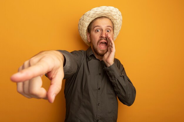 Surprised young handsome man in grey shirt and summer hat shouting or calling someone with hand near mouth and pointing with index finger at something 