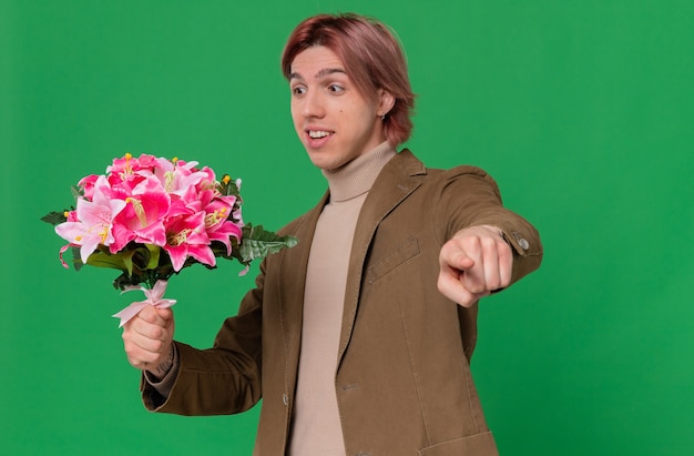 Surprised young handsome man holding and looking at bouquet of flowers pointing forward