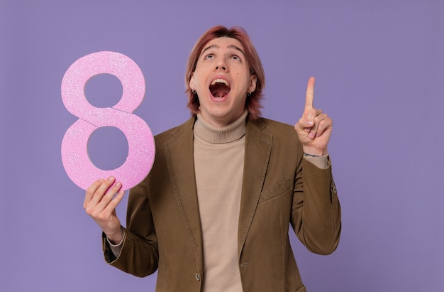 Free Photo surprised young handsome man holding pink number eight looking and pointing up 