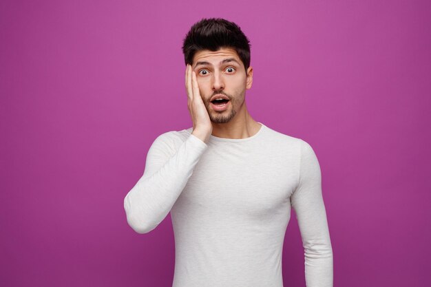 Surprised young handsome man looking at camera keeping hand on face on purple background