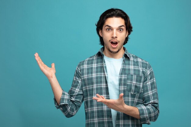 surprised young handsome man looking at camera showing empty hands isolated on blue background