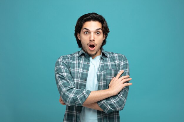 surprised young handsome man looking at camera while keeping arms crossed isolated on blue background