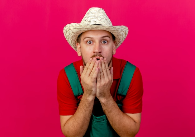 Free Photo surprised young handsome slavic gardener in uniform and hat looking keeping hands on mouth 