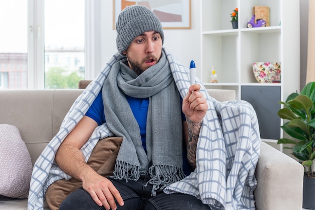 Surprised young ill man wearing scarf and winter hat wrapped in blanket sitting on sofa in living room keeping hand on leg holding and looking at thermometer