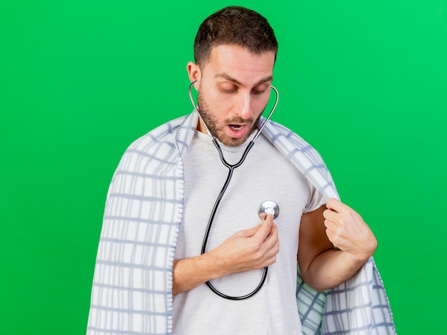 Free photo surprised young ill man wrapped in plaid wearing and listening to his own heartbeat with stethoscope isolated on green background