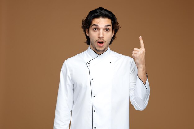 surprised young male chef wearing uniform looking at camera pointing up isolated on brown background