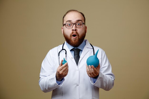 Surprised young male doctor wearing glasses lab coat and stethoscope around his neck holding enemas looking at camera isolated on olive green background