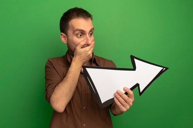 Free photo surprised young man holding and looking at arrow mark pointing at side keeping hand on mouth isolated on green wall