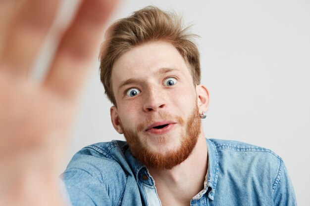 Surprised young man with beard in jean shirt stretching hand to camera.