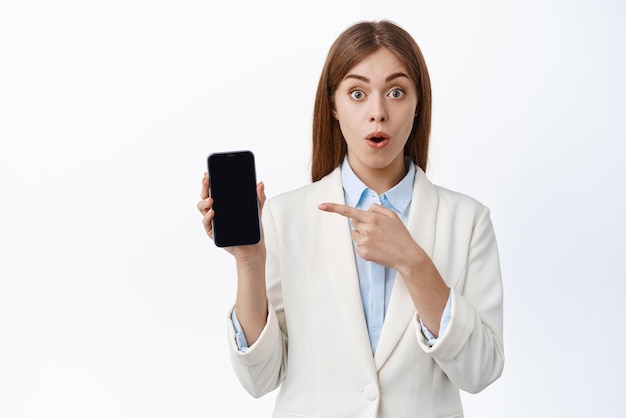 Surprised young office girl in business suit gasps and points left at smartphone screen shows online promo offer white background