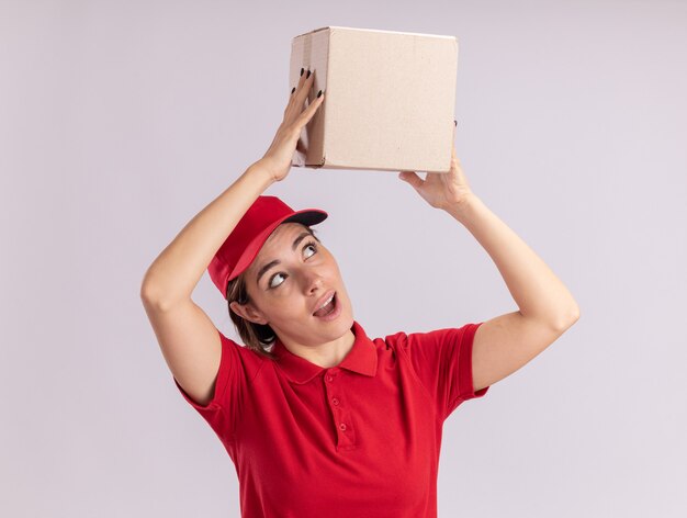 Surprised young pretty delivery girl in uniform holds cardbox over head on white