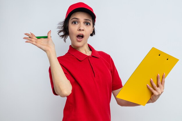 Surprised young pretty delivery woman holding clipboard and pen 