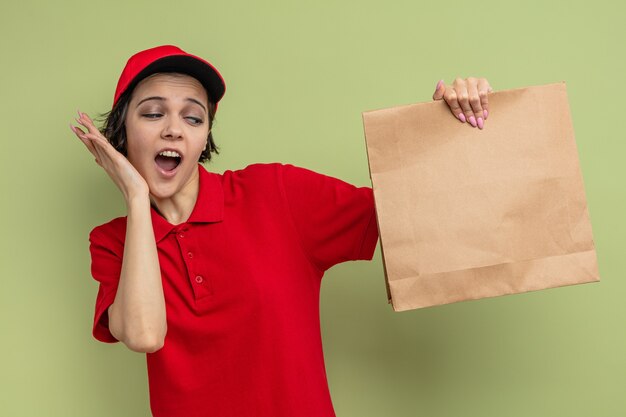 Surprised young pretty delivery woman putting hand on her face and holding paper food packaging 