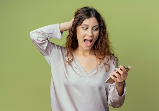 Surprised young pretty female office worker holding and looking at phone and putting hand on behind head isolated on olive green wall