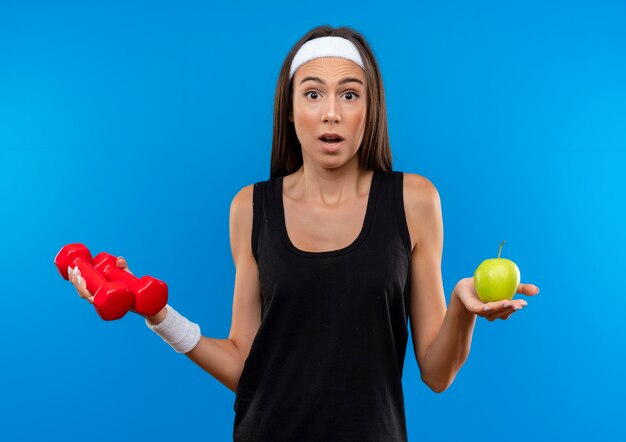 Surprised young pretty sporty girl wearing headband and wristband holding dumbbells and apple isolated on blue space