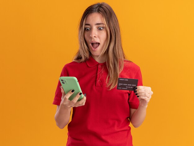 Surprised young pretty woman holds credit card and looks at phone isolated on orange wall