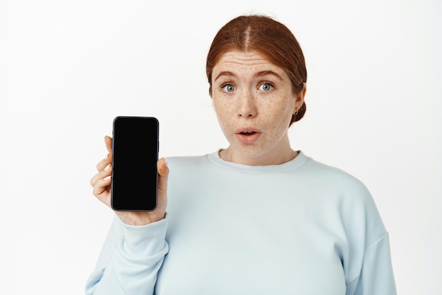 Surprised young redhead female showing smartphone mobile screen, raise eyebrows and gasp amazed, demonstrating cellphone app interface, white background.