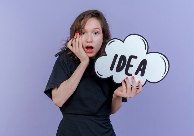 Surprised young slavic female barber wearing uniform holding scissors and idea bubble touching face isolated on purple background with copy space