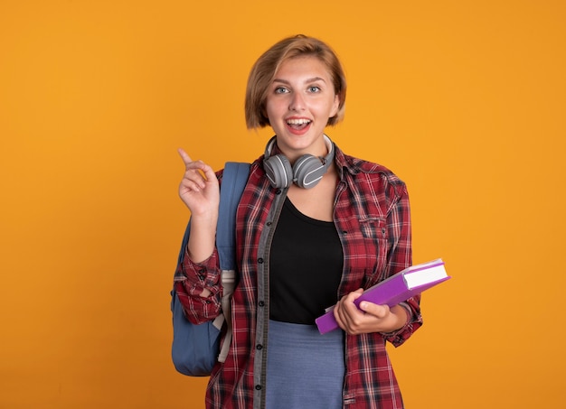 Surprised young slavic student girl with headphones wearing backpack holds book and notebook points at side 