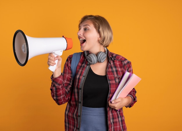 Free photo surprised young slavic student girl with headphones wearing backpack holds book and notebook shouts into loud speaker