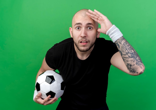 Free Photo surprised young sporty man wearing wristband holding ball and looking at distance with hand isolated on green