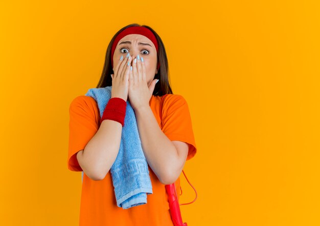 Surprised young sporty woman wearing headband and wristbands with towel and jump rope on shoulders  keeping hands on mouth isolated on orange wall with copy space