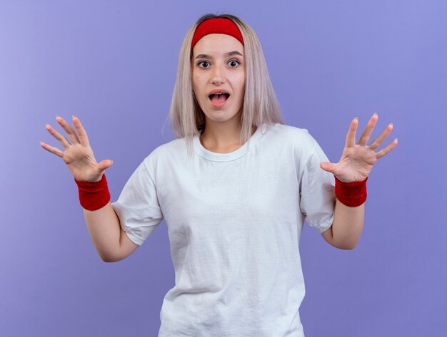 Surprised young sporty woman with braces wearing headband and wristbands holds hands open isolated on purple wall