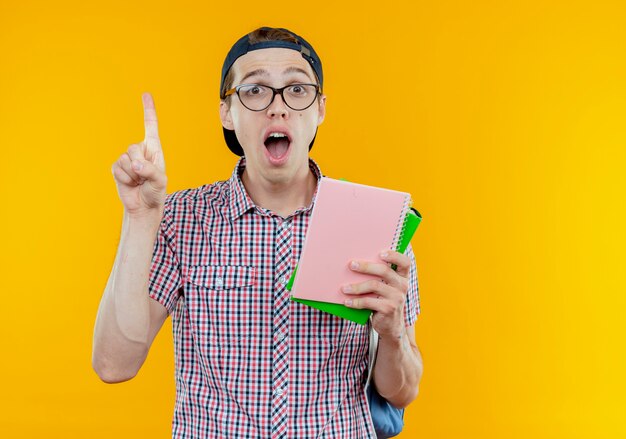 Surprised young student boy wearing back bag and glasses and cap holding notebook and points at up on white