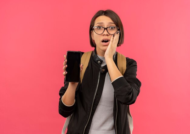 Surprised young student girl wearing glasses and back bag holding mobile phone putting hand on face isolated on pink wall