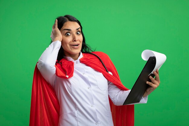 Surprised young superhero girl wearing medical robe with stethoscope holding clipboard putting hand on head isolated on green