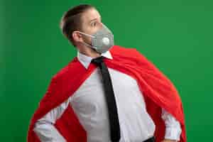 Free photo surprised young superhero guy wearing medical mask and tie looking at side isolated on green background