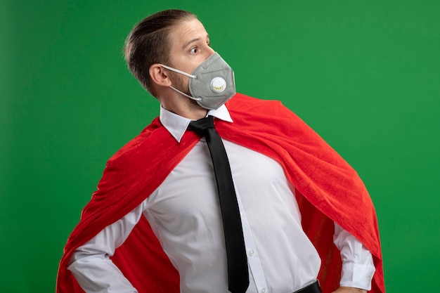 Free Photo surprised young superhero guy wearing medical mask and tie looking at side isolated on green background