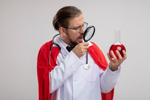 Free photo surprised young superhero guy wearing medical robe with stethoscope and glasses holding and looking at chemistry glass bottle filled with red liquid with magnifier isolated on white background