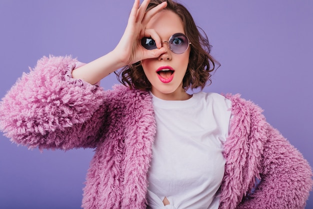 Free photo surprised young woman in fluffy jacket making faces on purple wall. spectacular white girl in sunglasses fooling around.