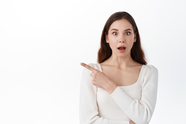 Surprised young woman look in awe pointing index finger aside showing awesome advertisement demonstrate banner wright your text here standing against white background