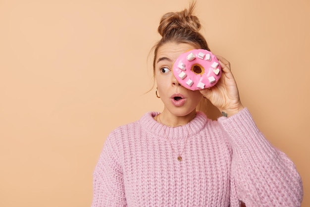 Free photo surprised young woman looks with shocked expression away keeps mouth opened covers eye with glazed doughnut going to eat delicious snack wears knitted sweater isolated over beige background.