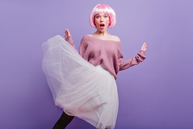 Free photo surprised young woman in lush white skirt posing on purple wall.  fashionable female model in pink peruke expressing amazement.