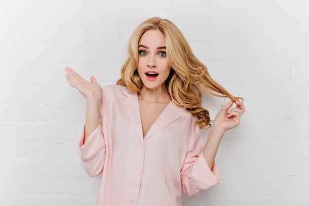 Surprised young woman in silk pyjamas plays with her wavy hair. Fair-haired girl in night-suit expressing amazement near bricked wall.