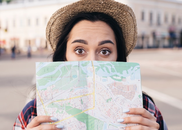 Free photo surprised young woman wearing hat covering her mouth with map at outdoors