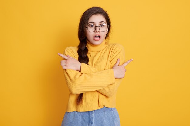 Surprised young woman wearing stylish clothing pointing with index fingers to sides