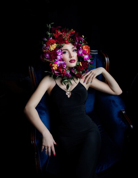 Surrounded face with colourful flowers, beautiful girl dressed in black dress on the blue armchair and bright make-up