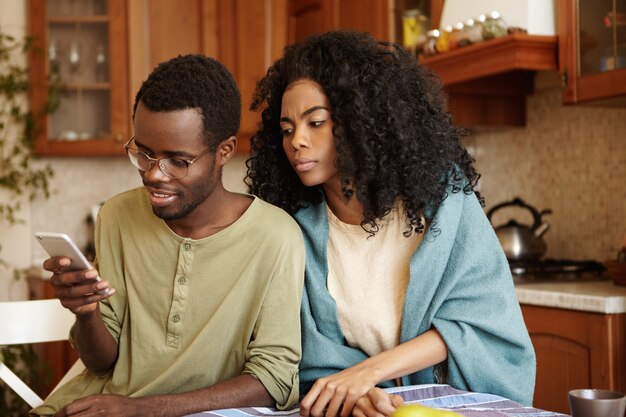 Suspicious black wife trying to read message that her happy husband sending to someone on mobile phone as she suspects betrayal, not trusting him. Jealousy, infidelity and distrust