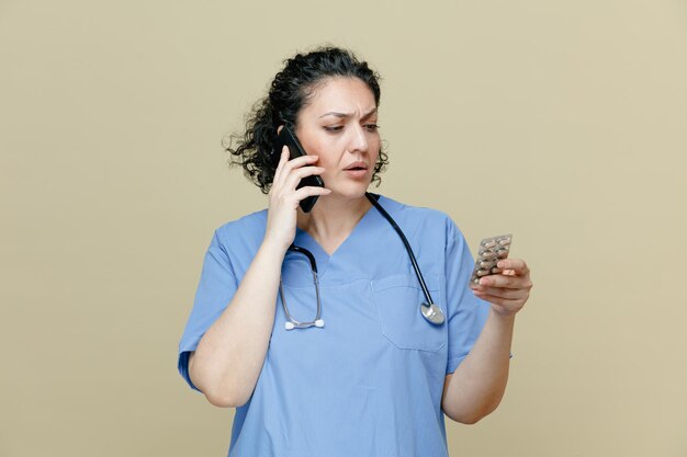 Suspicious middleaged female doctor wearing uniform and stethoscope around neck holding pack of pills talking on phone looking at pack isolated on olive background