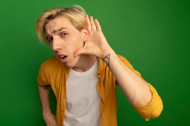 Free photo suspicious young blonde guy wearing yellow t-shirt showing listen gesture isolated on green