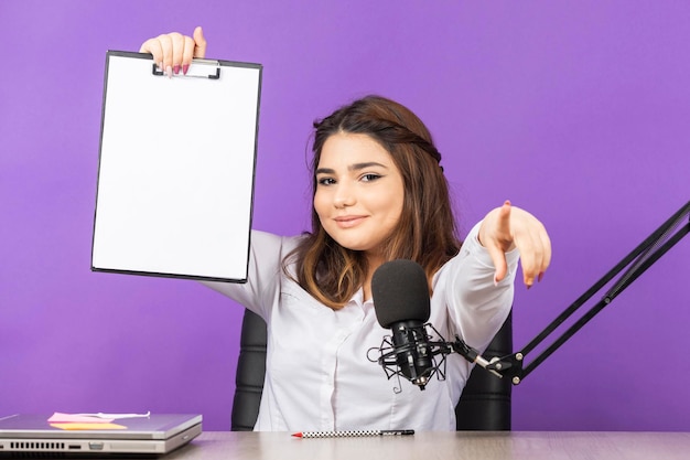 Sweet girl holding notebook and point finger to the camera Beautiful girl sitting behind desk on purple background High quality photo