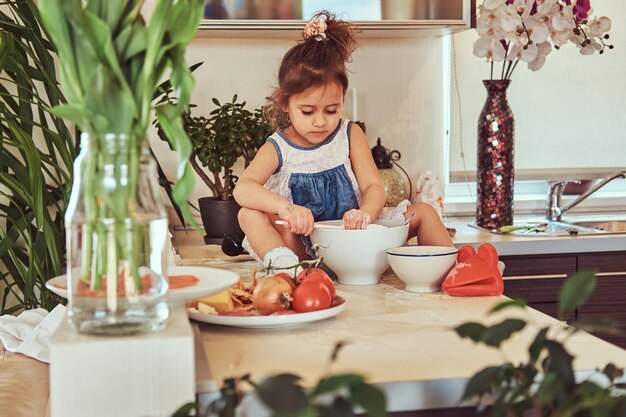 Sweet little cute girl learns to cook a meal in the kitchen while sitting on a countertop.