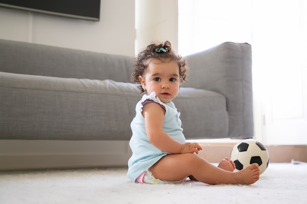 Sweet serious black haired baby girl in pale blue clothes sitting on floor with soccer ball, a. Side view. Kid at home and childhood concept