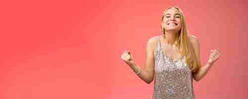 Free photo sweet taste win charming happy blond girl rejoicing triumphing raise head up close eyes smiling broadly clench fists victory celebration gesture achieve dream come true standing red background