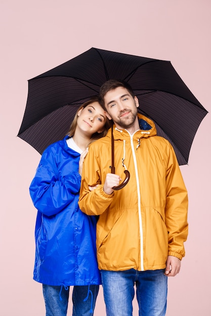 Sweet young beautiful couple posing in rain coats holding umbrella over light pink wall