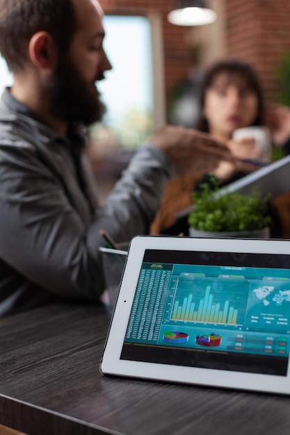 Tablet computer with company statistics on screen standing on table in startup office. Millennial multiethnic businesspeople discussing marketing strategy working at business collaboration